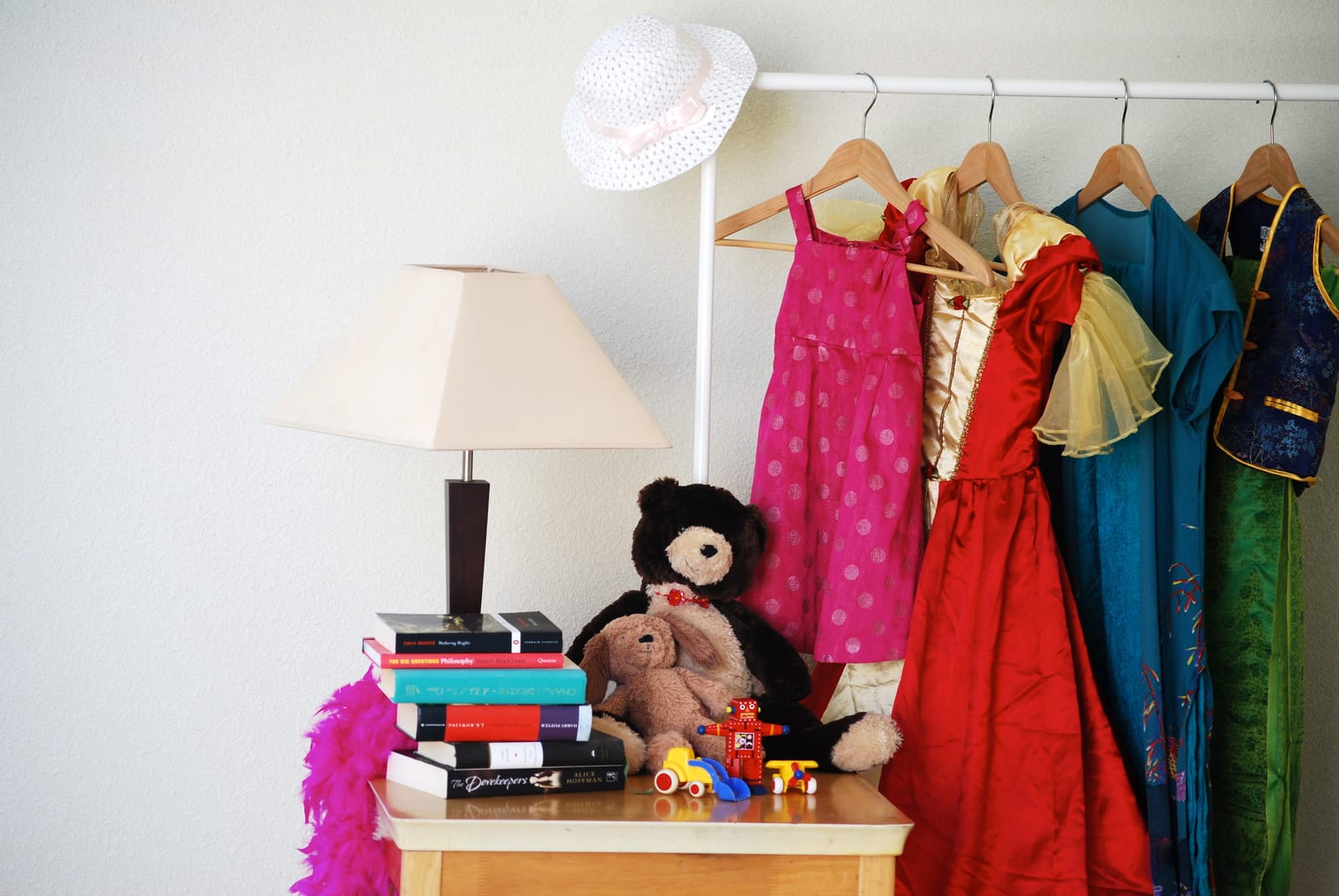 A yard sale: books, toys, a lamp and a hat on a small table beside a rail of dresses.
